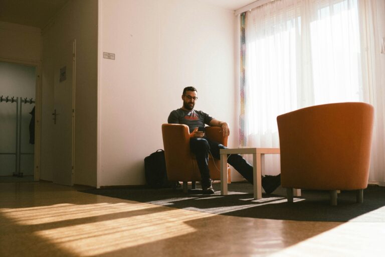A man sits in a modern lounge, enjoying sunlight through large windows.
