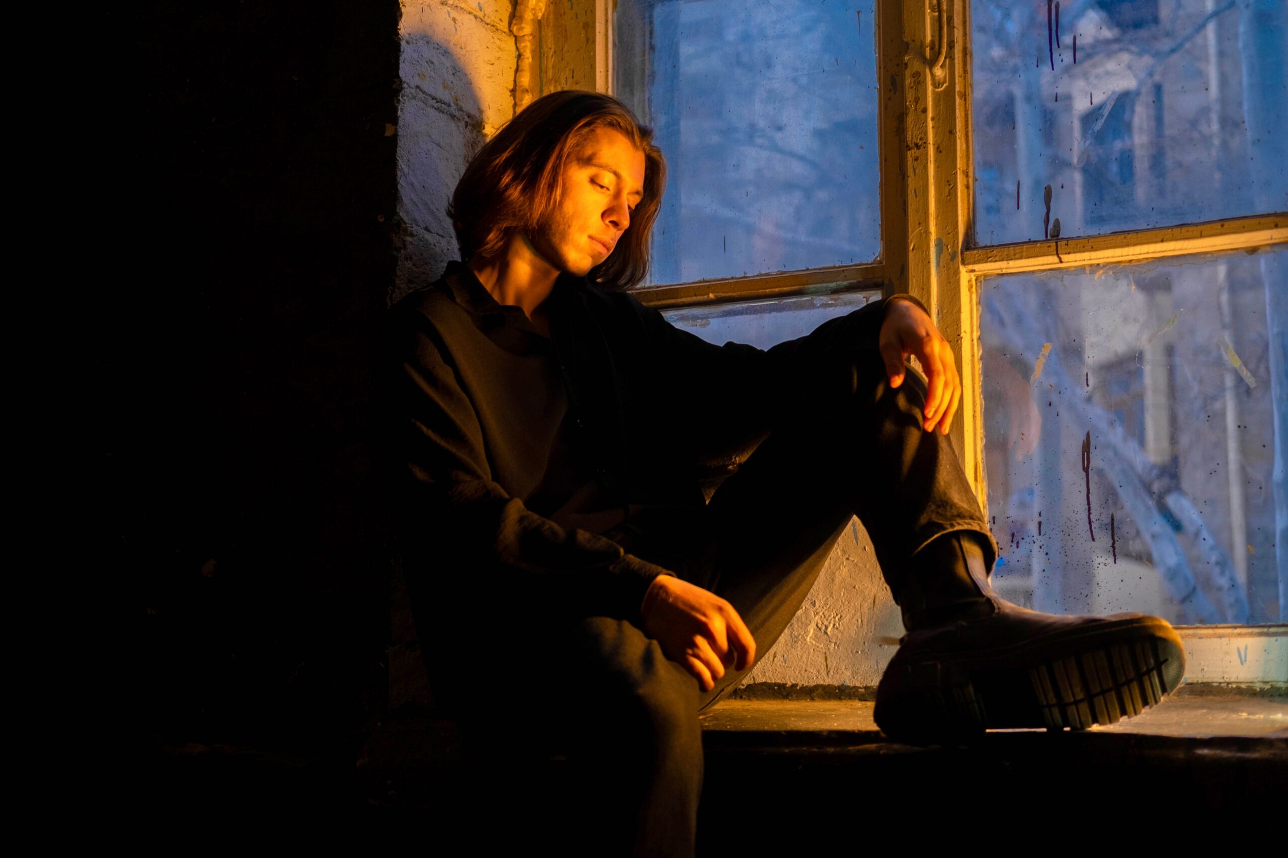 A contemplative young adult sits by a window, illuminated by warm evening light.